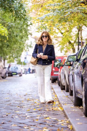 Téléchargez les photos : Une femme aux cheveux blonds marche dans la rue et prend un café. Jolie femme portant un manteau bleu et un pantalon blanc. - en image libre de droit