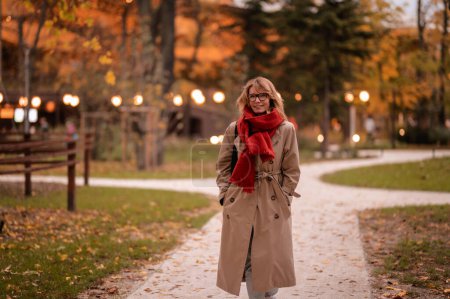 Photo for Woman walking in the public park in autumn. Blond haired female wearing scarf and trench coat. - Royalty Free Image