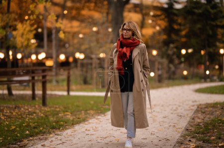 Téléchargez les photos : Toute la longueur d'une femme marchant dans le parc public en automne. Femme aux cheveux blonds portant écharpe et trench coat. - en image libre de droit