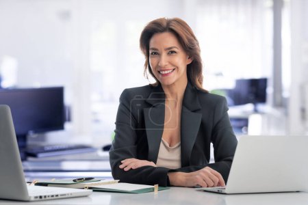 Foto de Morena de pelo de mediana edad empresaria sentada en la oficina y el uso de computadoras portátiles para el trabajo. Mujer profesional de confianza que usa casual de negocios. - Imagen libre de derechos