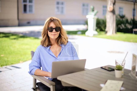 Foto de Una rubia de mediana edad sentada en la terraza de un café y usando un portátil para trabajar. Mujer confiada mirando a la cámara y sonriendo. - Imagen libre de derechos