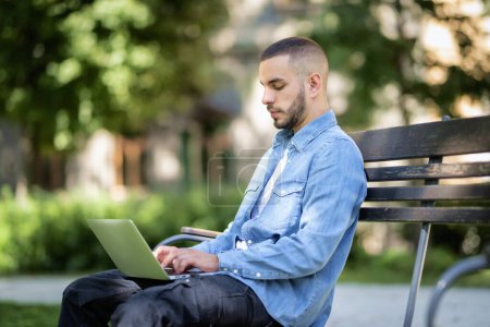Téléchargez les photos : Beau jeune homme assis sur un banc dans un parc public et utilisant un ordinateur portable. Il porte des vêtements décontractés et travaille ou apprend sur Internet. - en image libre de droit