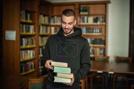 Photo for Young man standing in the university library and holding books in his hand. - Royalty Free Image