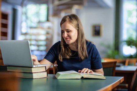 Téléchargez les photos : Une femme malvoyante assise et étudiant à la bibliothèque de l'université. Jeune femme utilisant un ordinateur portable et des livres. - en image libre de droit