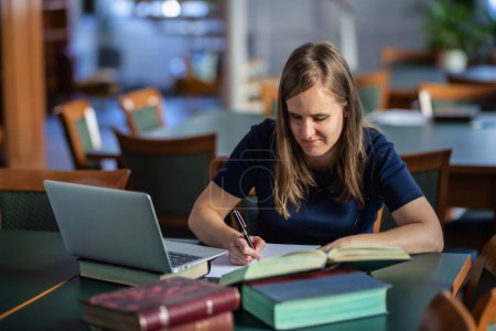 Téléchargez les photos : Une femme malvoyante assise et étudiant à la bibliothèque de l'université. Jeune femme utilisant un ordinateur portable et des livres. - en image libre de droit