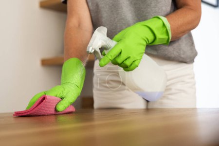 Photo for Close-up of a woman's hand wiping the dining table at home. Confident female wearing rubber gloves and using cleaning liquid while cleaning at home. - Royalty Free Image