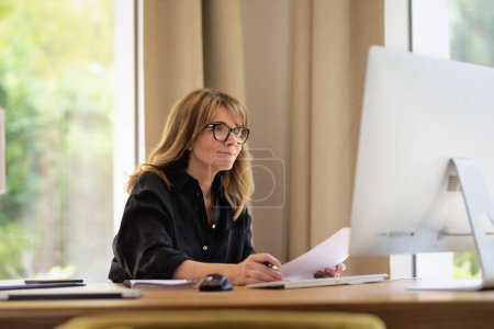 Téléchargez les photos : Femme d'âge moyen assis au bureau tenant un document dans sa main et utilisant l'ordinateur pour le travail. Femme d'affaires confiante travaillant à la maison. Bureau à domicile. - en image libre de droit