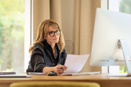 Photo for Middle aged woman sitting at desk holding a document in her hand and using computer for work. Confident businesswoman working at home. Home office. - Royalty Free Image