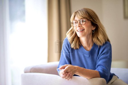 Foto de Primer plano de una mujer de mediana edad con el pelo rubio está sentado en un sillón en casa. Mujer segura vistiendo camisa azul y gafas y sonriente alegre. - Imagen libre de derechos