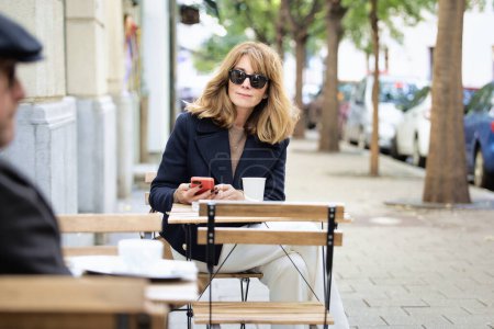 Téléchargez les photos : Une femme d'âge moyen assise à une table devant un café et utilisant son smartphone tout en buvant une tasse de café. Femme confiante portant des vêtements décontractés et des lunettes de soleil. - en image libre de droit