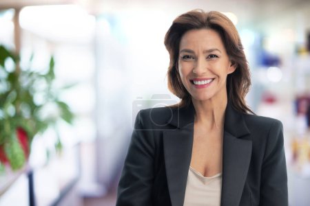 Téléchargez les photos : Femme d'affaires d'âge moyen confiante portant un blazer noir et souriant au bureau. - en image libre de droit