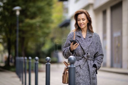 Photo for Attractive woman walking on the street and using smartphone. Middle aged female wearing winter coat and text messaging. - Royalty Free Image