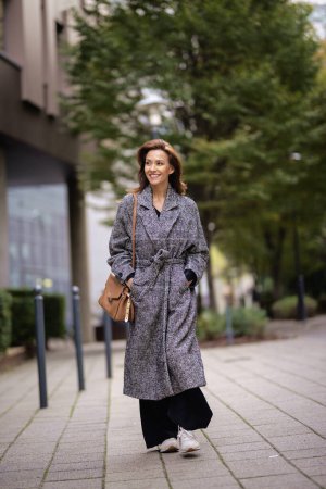 Photo for Full length of a middle adult woman with brunette hair walking outdoors in city street on autumn day. She is wearing tweed coat. - Royalty Free Image