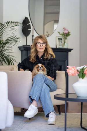 Foto de Atractiva mujer de pelo rubio con su lindo cachorro sentado en un sillón en una casa moderna. Mujer sonriente vistiendo camisa negra y jeans. - Imagen libre de derechos