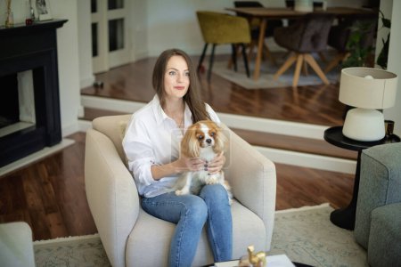 Photo for Portrait of beautiful long haired woman with her cute puppy relaxing in an armchair in her modern home. home. - Royalty Free Image