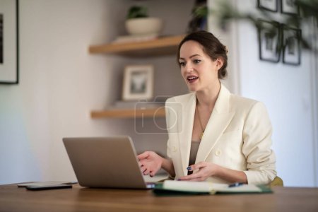 Téléchargez les photos : Jolie jeune femme d'affaires avec ordinateur portable assis au bureau et ayant web conférence au bureau à domicile. - en image libre de droit