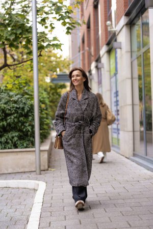 Photo for Full length of a middle adult woman with brunette hair walking outdoors in city street on autumn day. She is wearing tweed coat. - Royalty Free Image