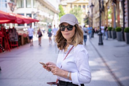 Photo for Attractive woman walking on sidewalk in the city and text messaging. Middle aged female wearing white shirt and black pants. Full length shot. - Royalty Free Image