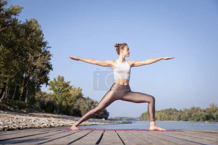 Téléchargez les photos : Toute la longueur de la femme caucasienne pratiquant le yoga sur la jetée près de la rivière. Attrayant femelle a les cheveux blonds et souriant. Guerrier 2 pose de yoga. - en image libre de droit