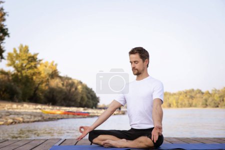 Foto de Hombre de mediana edad practicando yoga al aire libre. Hombre caucásico usando esterilla de yoga y haciendo pose de loto sobre la naturaleza. - Imagen libre de derechos