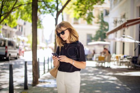Photo for Attractive woman with blonde hair walking on the street and using a mobile phone. Cheerful smiling female wearing sunglasses and text messaging. - Royalty Free Image