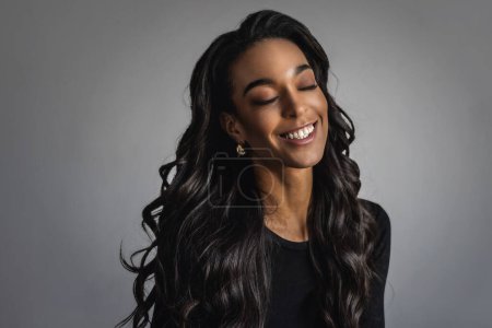 Foto de Alegre retrato femenino afroamericano sonriente. Mujer negra de pelo largo con los ojos cerrados sobre fondo gris. Copiar espacio. - Imagen libre de derechos