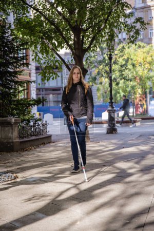 Foto de Mujer ciega con bastón blanco caminando por la ciudad. Una mujer con discapacidad visual que viste ropa casual y usa su bastón para cruzar la calle. - Imagen libre de derechos