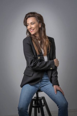 Foto de Atractiva mujer de mediana edad con sonrisa dentada usando chaqueta negra y jeans mientras se sienta en un fondo gris aislado. Copiar espacio. Captura de estudio. - Imagen libre de derechos