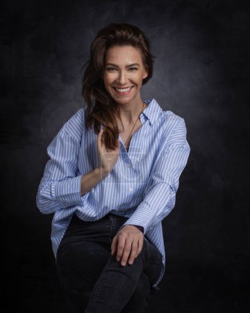 Foto de Atractiva mujer de mediana edad con sonrisa dentada usando camisa azul y jeans negros mientras se sienta en un fondo oscuro aislado. Copiar espacio. Captura de estudio. - Imagen libre de derechos