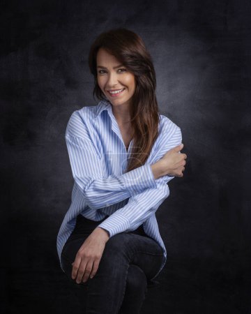 Photo for Attractive middle aged woman with toothy smile wearing blue shirt and black jeans while sitting against at isolated dark background. Copy space. Studio shot. - Royalty Free Image