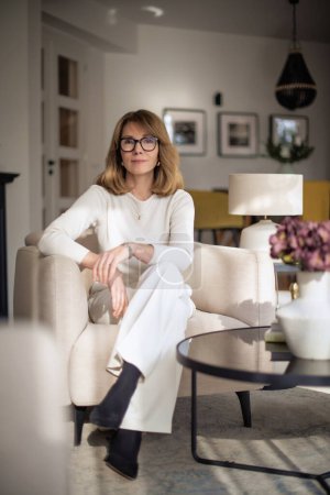 Photo for Attractive middle aged woman relaxing in an armchair at home. Blond haired female wearing eyeglasses and white sweater and pants. Full length shot. - Royalty Free Image