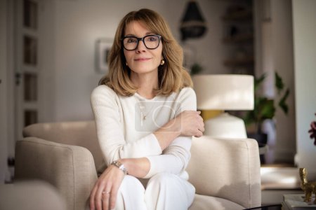 Photo for Close-up of an attractive middle aged woman relaxing in an armchair at home. Blond haired female wearing eyeglasses and white sweater and pants. - Royalty Free Image