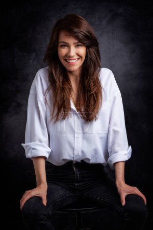 Photo for Beautiful smiling woman with white teeth sitting at isolated dark grey background. Happy woman wearing white shirt and black jeans. Copy space. - Royalty Free Image