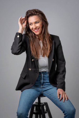 Photo for Beautiful smiling woman with white teeth sitting at isolated grey background. Happy woman wearing black jacket and jeans. Studio shot. Copy space. - Royalty Free Image