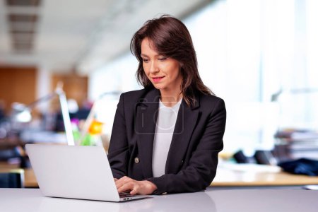 Foto de Atractiva mujer de negocios con chaqueta y el uso de la computadora portátil para el trabajo. Mujer sonriente sentada en la oficina y trabajando. - Imagen libre de derechos