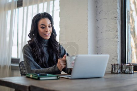 Photo for An African-American woman sitting at the table by the window and working on her laptop. Home office. Attractive  woman  having web conference. - Royalty Free Image