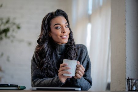 Foto de Una mujer afroamericana sentada en la mesa junto a la ventana y bebiendo té. Oficina central. Mujer atractiva con ropa casual - Imagen libre de derechos