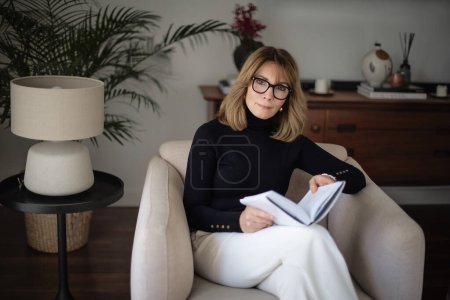 Téléchargez les photos : Photo d'une femme d'âge moyen lisant un livre dans son salon pendant les vacances d'hiver. Attrayant féminin portant un pull noir et des lunettes. - en image libre de droit