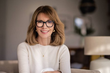 Photo for Close-up of an attractive middle aged woman relaxing in an armchair at home. Blond haired female wearing eyeglasses and white sweater. - Royalty Free Image