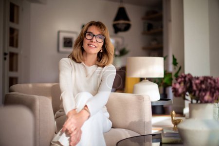 Photo for Attractive middle aged woman relaxing in an armchair at home. Blond haired female wearing eyeglasses and white sweater and pants. - Royalty Free Image