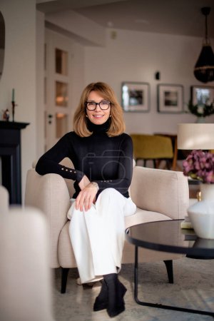 Photo for Attractive middle aged woman relaxing in an armchair at home. Blond haired female wearing eyeglasses and black sweater and white pants. Full length shot. - Royalty Free Image