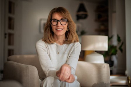 Photo for Attractive mid aged woman relaxing in an armchair at home. Blond haired female wearing eyeglasses and white sweater and pants. - Royalty Free Image