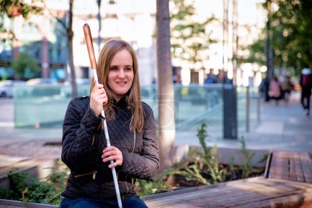 Photo for Close-up of a visually impaired woman holding a white cane and sitting on a bench in the city. Smiling female wearing casual clothes. - Royalty Free Image