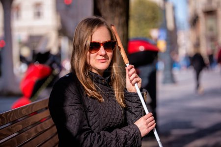 Foto de Primer plano de una mujer con discapacidad visual sosteniendo un bastón blanco y sentada en un banco de la ciudad. Mujer sonriente que usa ropa casual y gafas de sol. - Imagen libre de derechos
