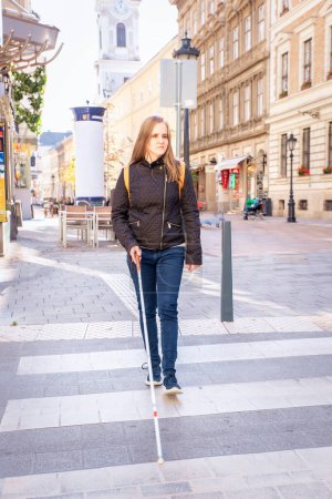 Photo for Portrait of blind woman with white cane crossing on the road in the city. A visually impaired woman wearing casual clothes and using her cane to cross the street. Full length shot. - Royalty Free Image