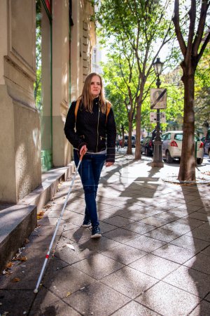 Foto de Retrato de mujer ciega con bastón blanco caminando por la calle. Una mujer con discapacidad visual que viste ropa casual y usa su bastón para caminar por la calle. Disparo completo. - Imagen libre de derechos