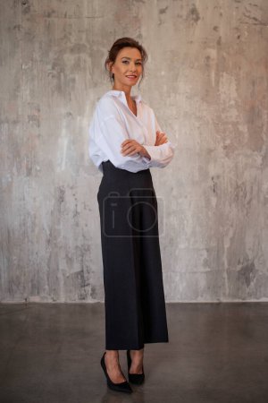 Téléchargez les photos : Une belle femme aux cheveux bruns debout devant un mur gris. Joyeux sourire féminin portant une chemise blanche et un pantalon noir et regardant la caméra. Plan complet. Espace de copie. - en image libre de droit