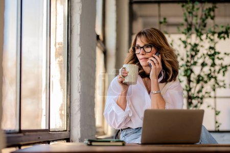 Photo for A middle-aged blonde woman with a mug in her hand looking thoughtfully and making a call. Attractive female wearing white shirt and smiling. Home office. - Royalty Free Image
