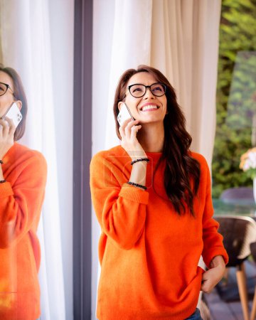 Photo for Attractive brunette haired woman standing by window and using her smartphone. Brunette haired female wearing orange sweater and talking with somebody on the phone. Candid shot. - Royalty Free Image