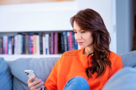 Photo for Attractive and happy woman using her smartphone and text messaging. Mid aged woman relaxing on the couch at home. She is wearing orange sweater and jeans. - Royalty Free Image
